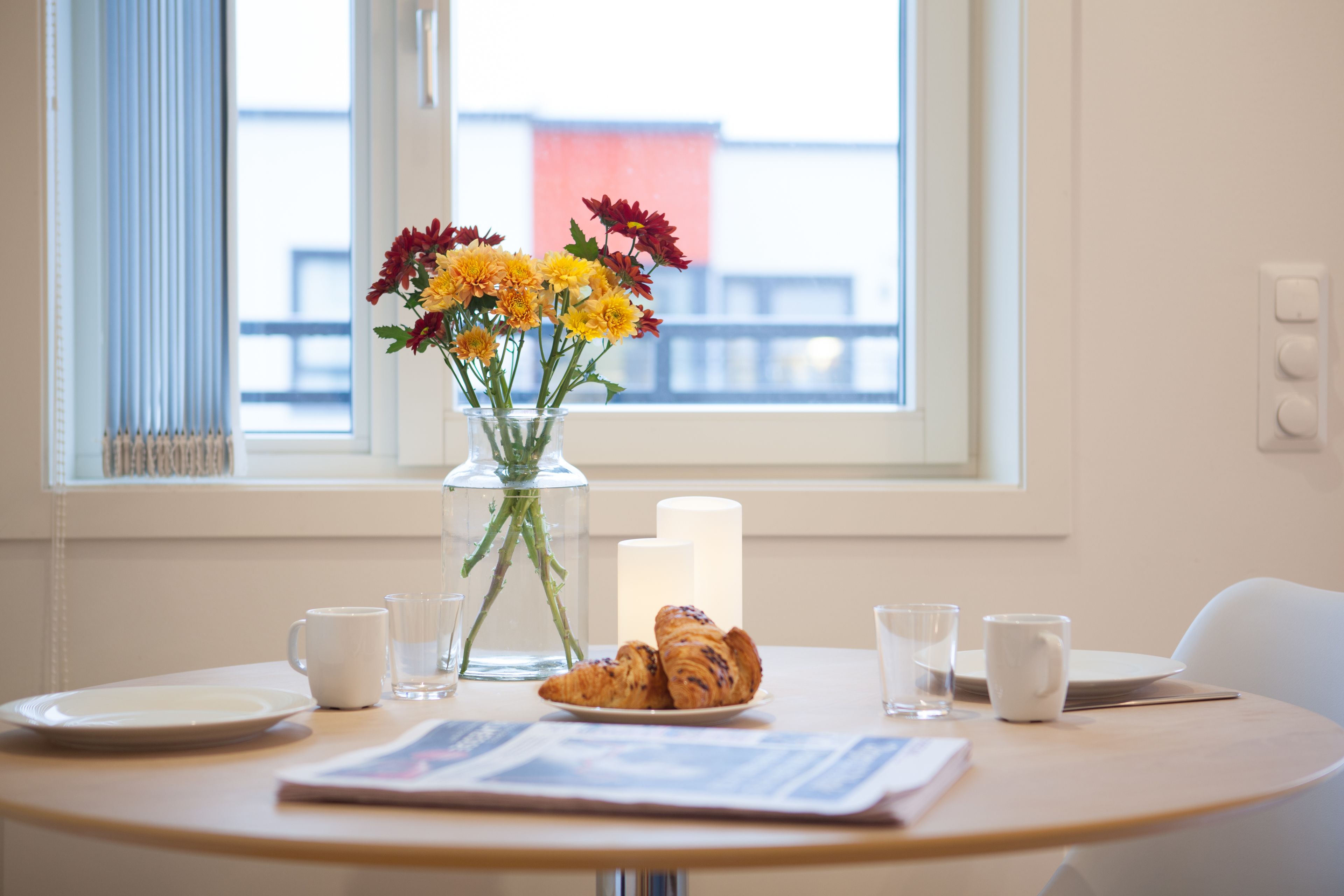 Une table avec un vase de fleurs, une bougie, des croissants et un journal.