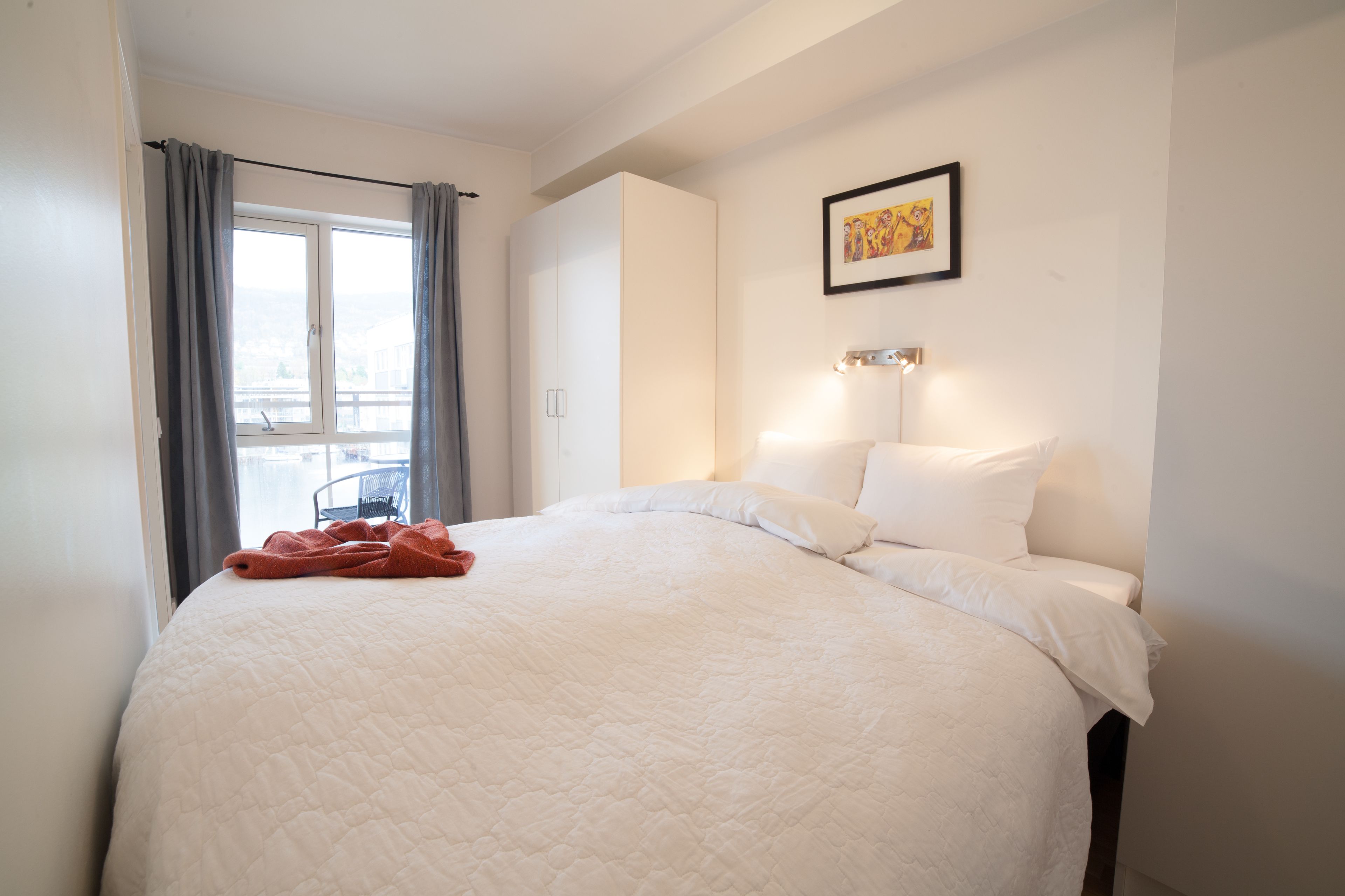 A neatly made bed with a white quilt, a red blanket, and a window with grey curtains.