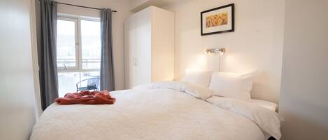 A neatly made bed with a white quilt, a red blanket, and a window with grey curtains.