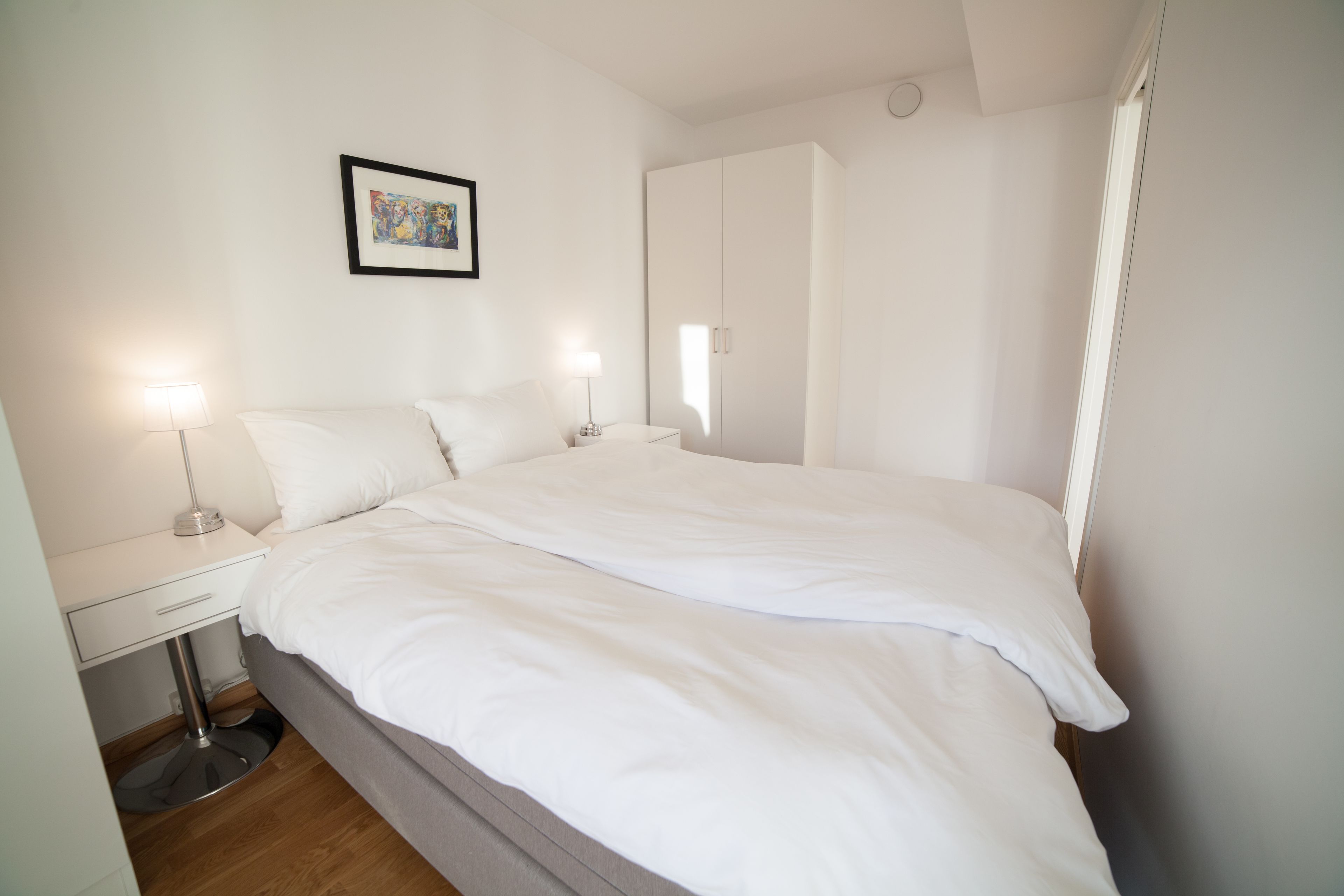 A neatly made bed with white linens, a bedside table with a lamp, and a framed picture on the wall.