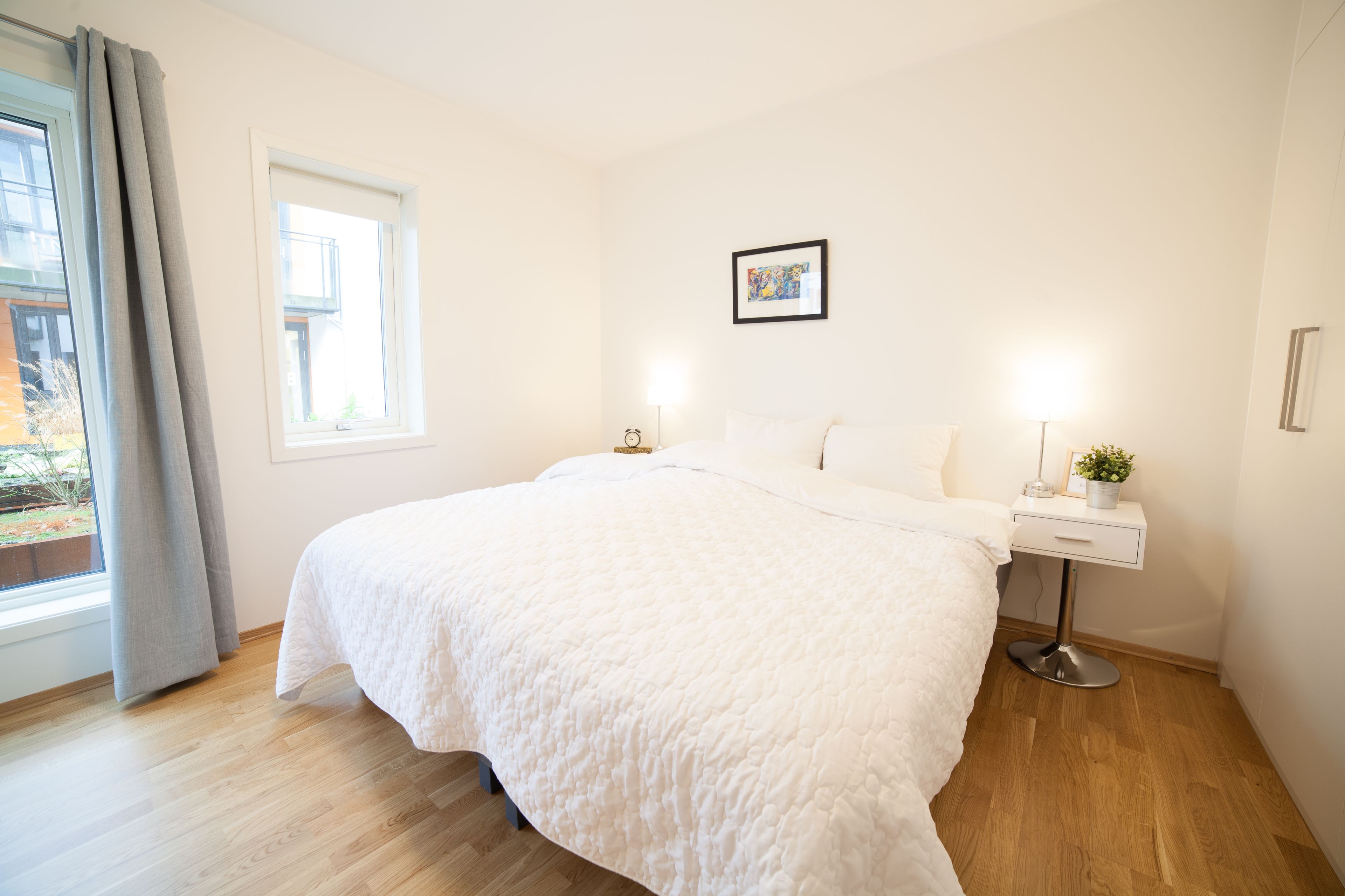 A bedroom with a large bed, a bedside table with a lamp, a window with curtains, and a framed picture on the wall.