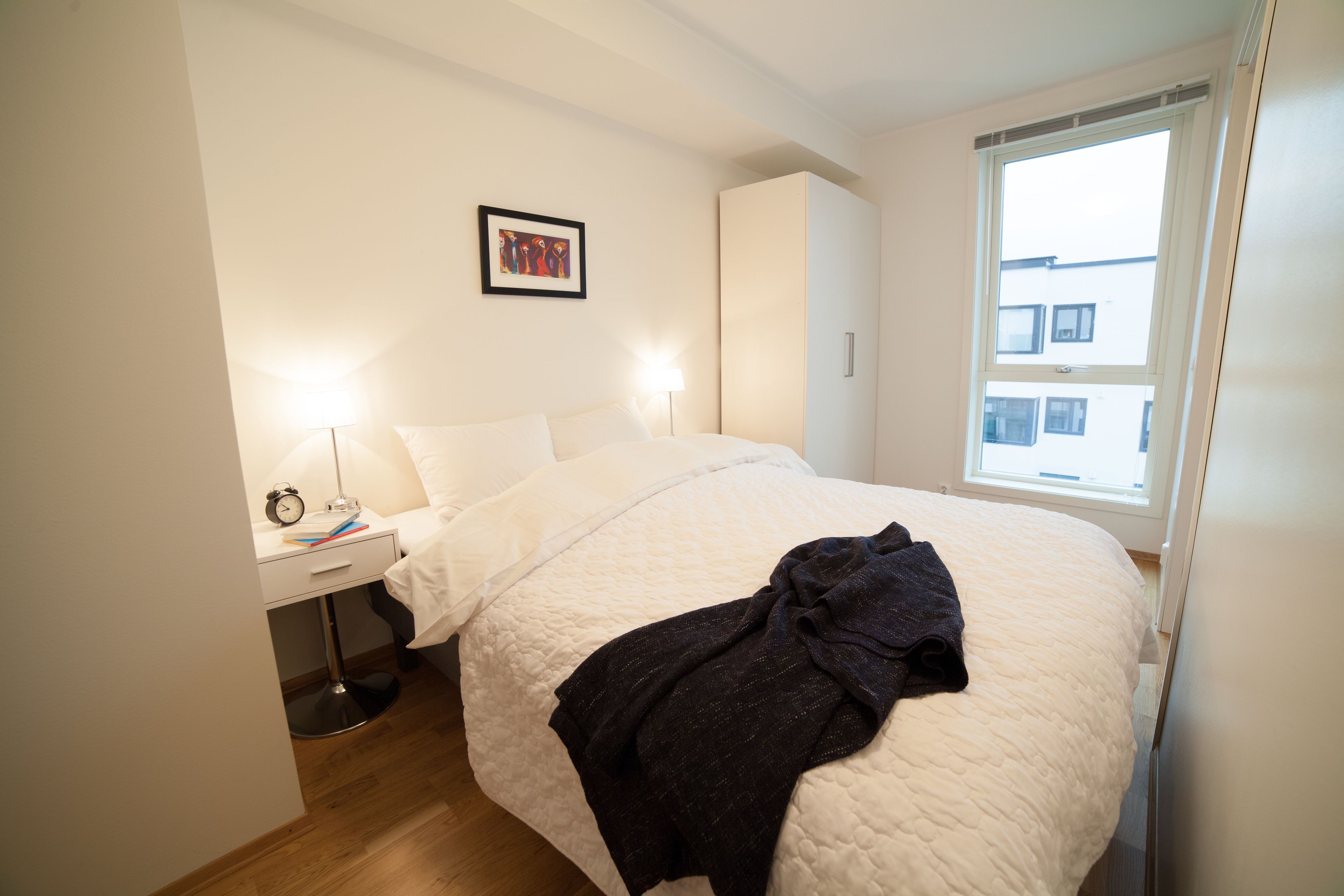 Une chambre à coucher bien rangée, avec un lit, une table de chevet, une lampe et une fenêtre donnant sur des immeubles.