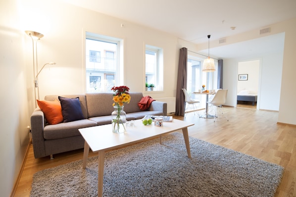 A modern living room with a grey sofa, a white coffee table, and a grey rug. There are windows with curtains, a floor lamp, and a dining area with a chair and table. - Bjørvika Apartments, Damsgård Area, Bergen city center (Bergen)
