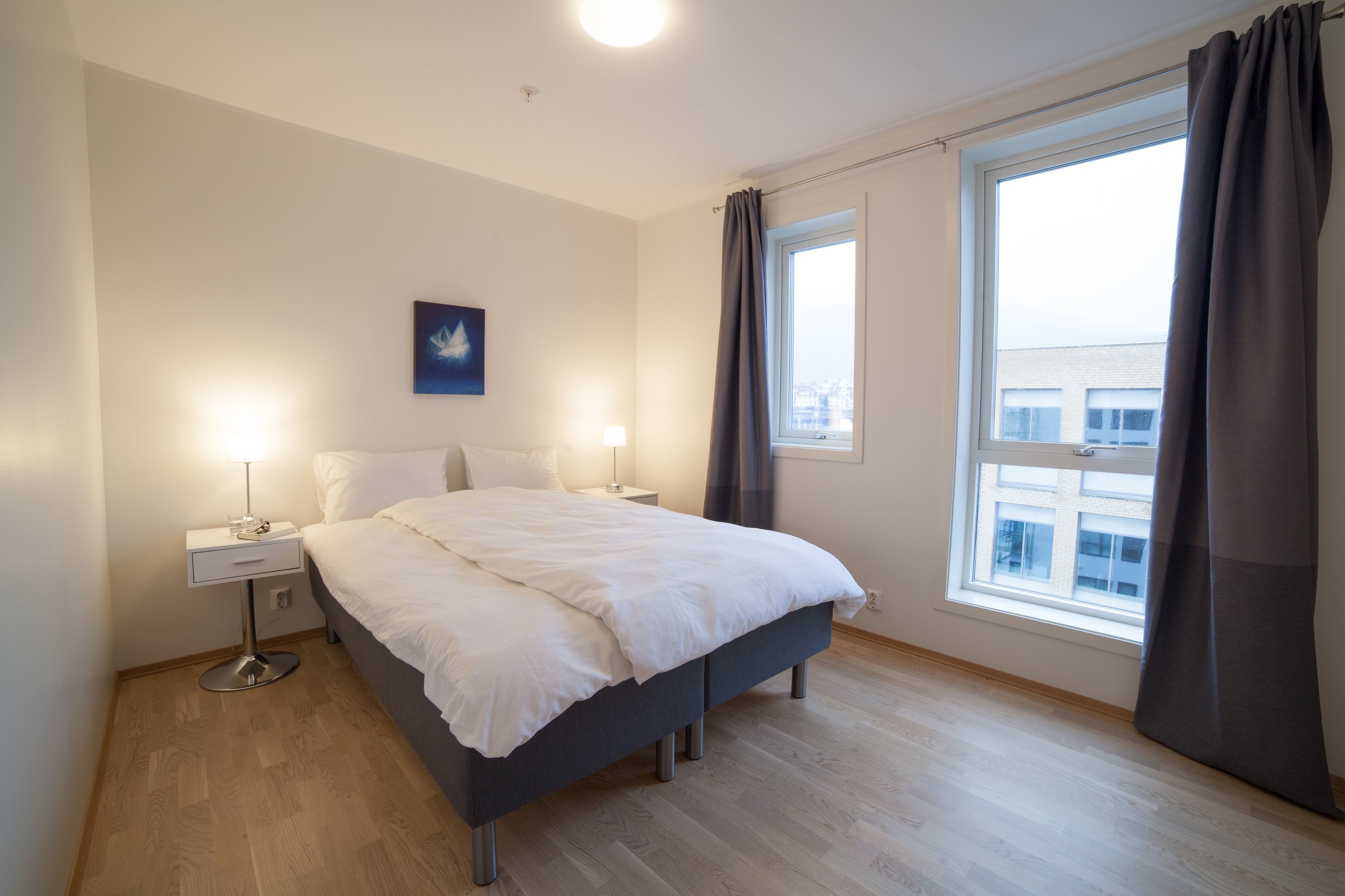 A modern bedroom with a large bed, two bedside tables, a nightstand, a window with curtains, and a city view.