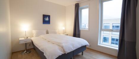 A modern bedroom with a large bed, two bedside tables, a nightstand, a window with curtains, and a city view.