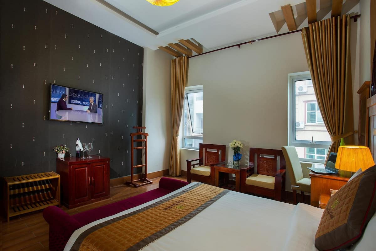 Habitación de hotel con una cama grande, un televisor mostrando noticias, un escritorio con una computadora portátil y una ventana con cortinas.