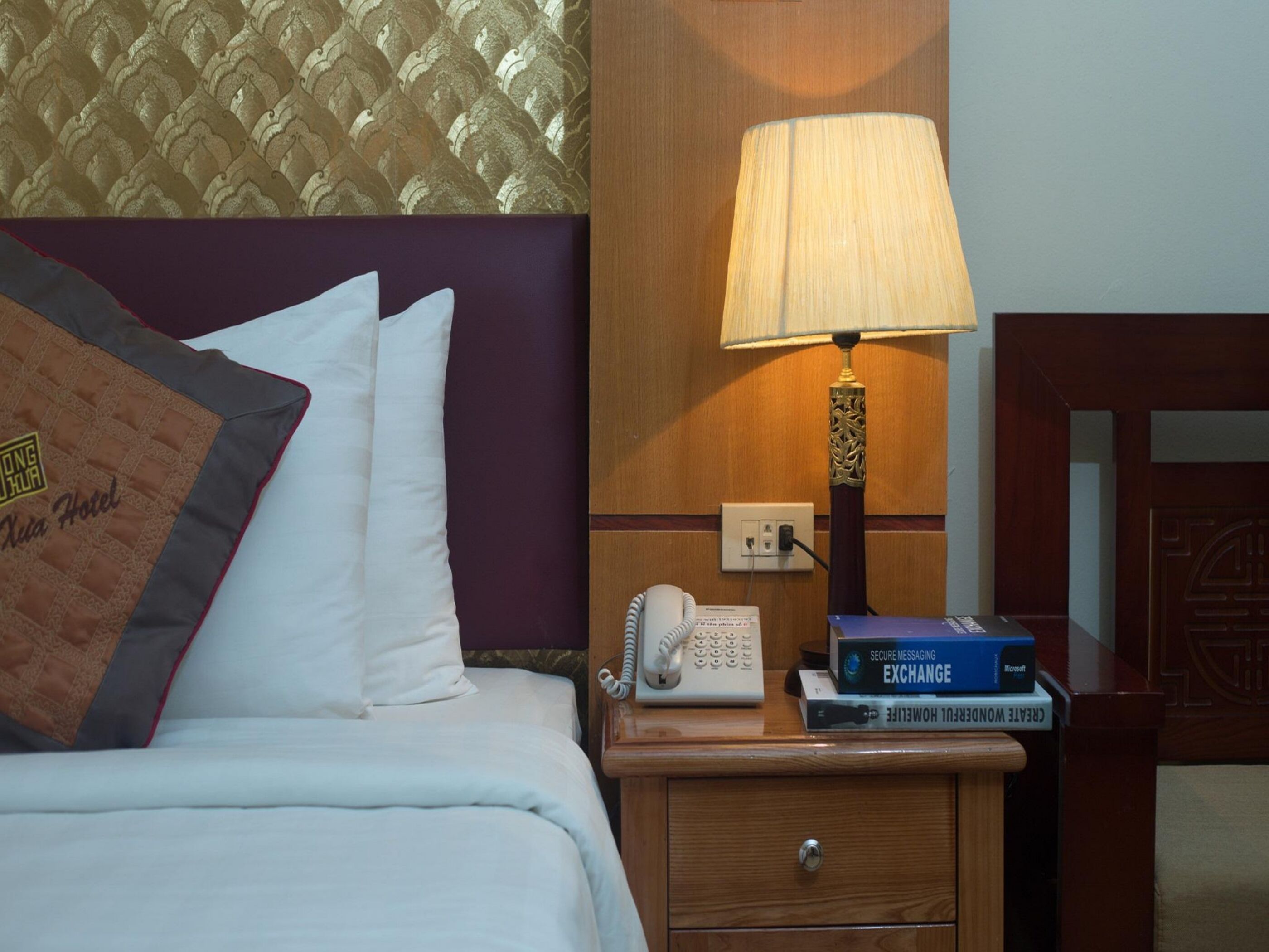 Quarto de hotel com uma cama, almofadas, mesa de cabeceira com abajur, telefone e alguns livros.