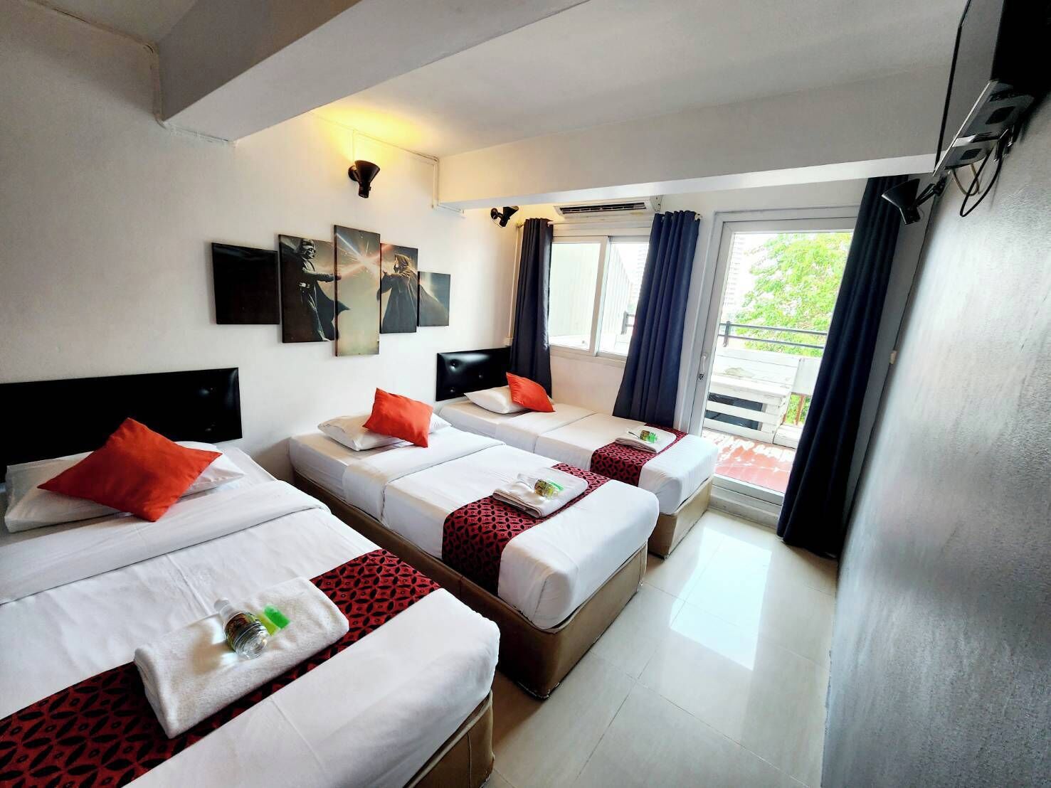 Une chambre d’hôtel avec trois lits simples, chacun agrémenté de draps blancs et de touches de couleur rouge. Les murs sont décorés d’œuvres d’art et il y a une fenêtre avec des rideaux.
