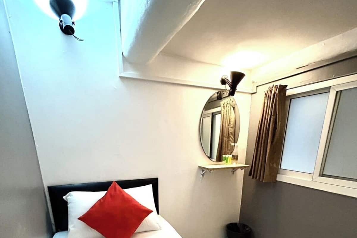 Une petite pièce, de taille modeste, comprenant un lit simple, un miroir rond, une fenêtre avec des rideaux et une petite poubelle.