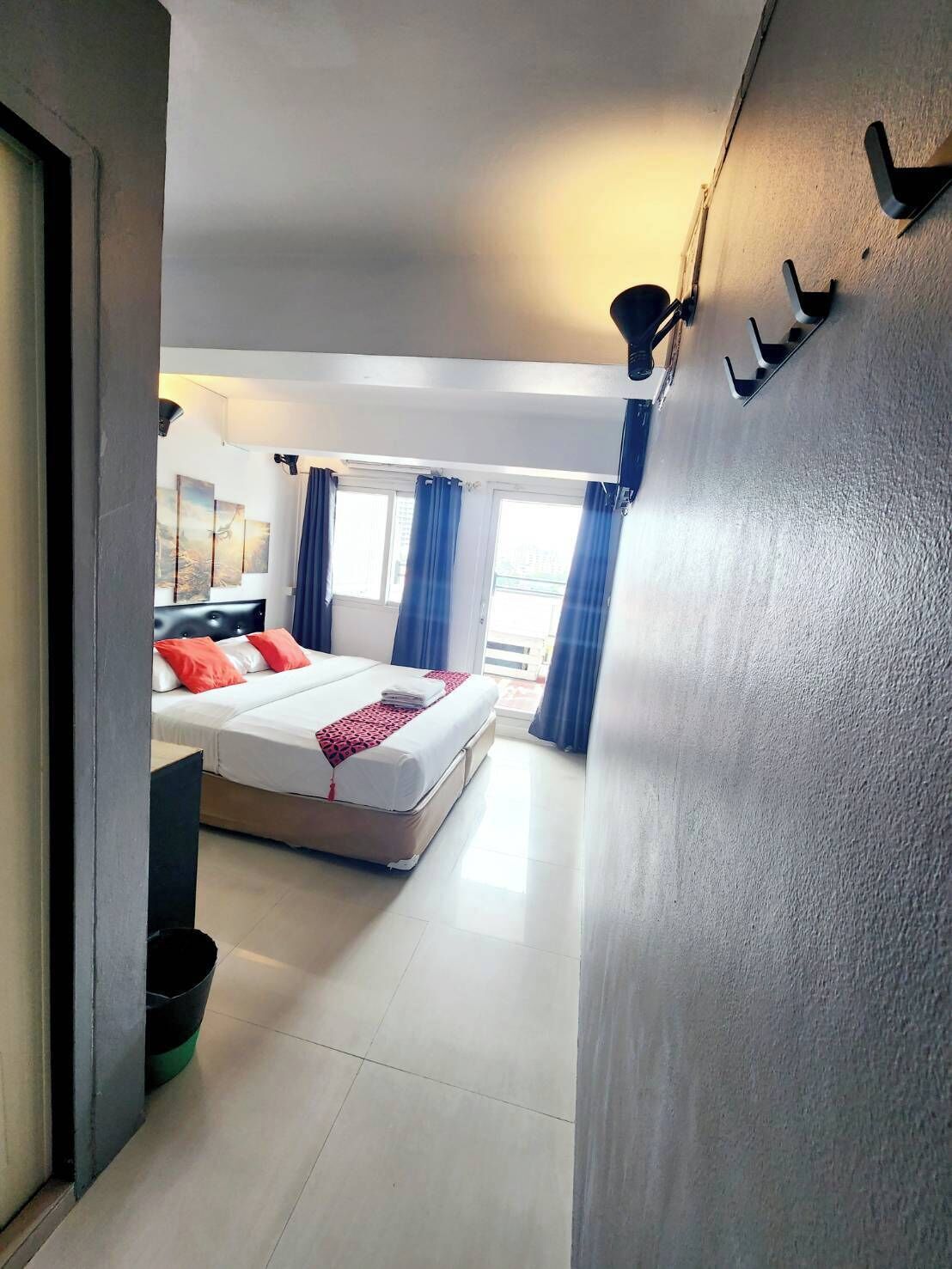 A hotel room with a bed, a window with curtains, a wall-mounted light, and a door.