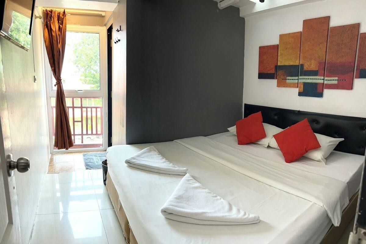 Une chambre d’hôtel avec un lit, du linge de lit blanc, des oreillers rouges, un mur foncé et une fenêtre avec des rideaux.