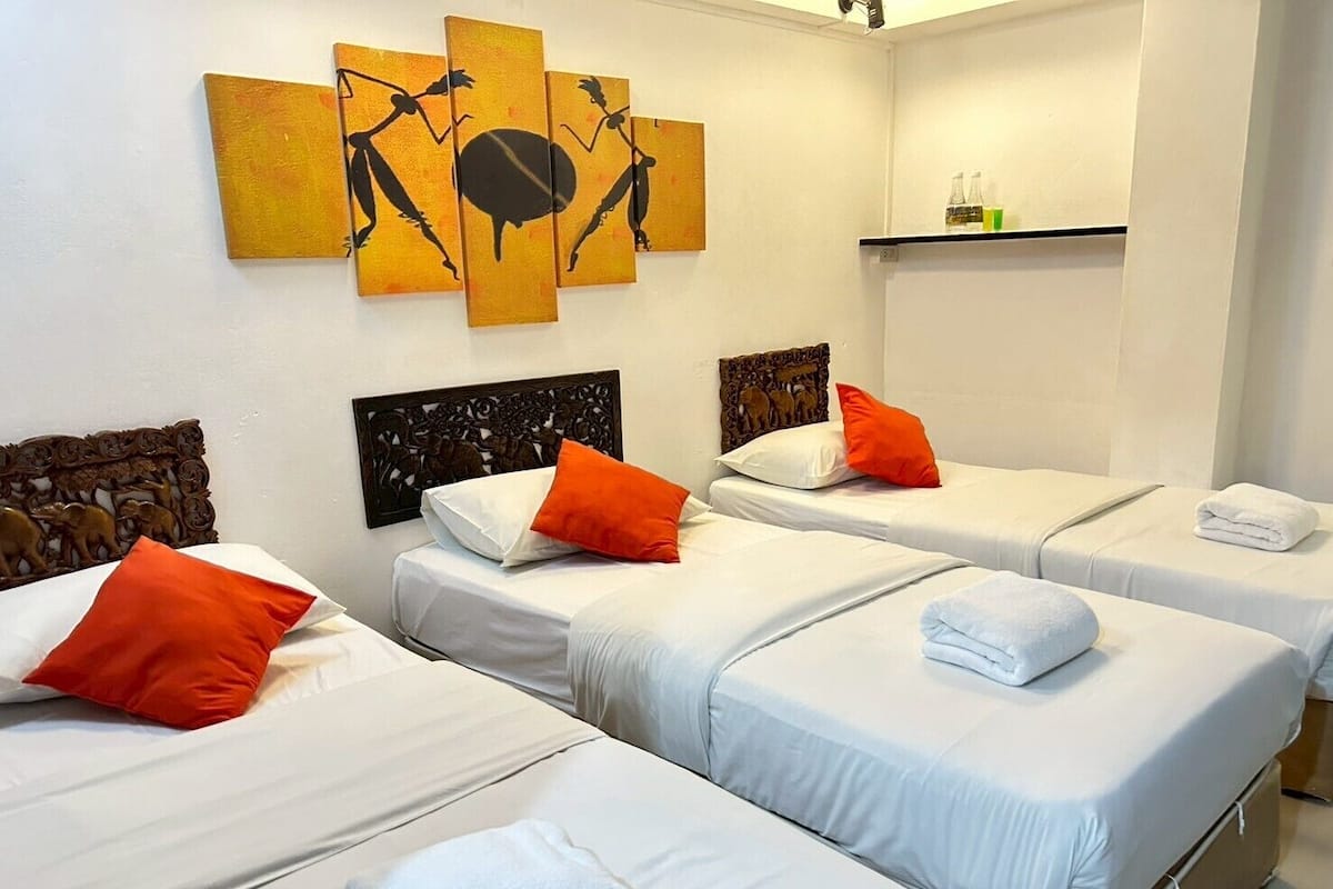 Une chambre d’hôtel avec trois lits, chacun agrémenté de draps blancs et d’oreillers orange. Un tableau décoratif est accroché au mur au-dessus des lits, et une étagère avec des bouteilles est fixée au mur.