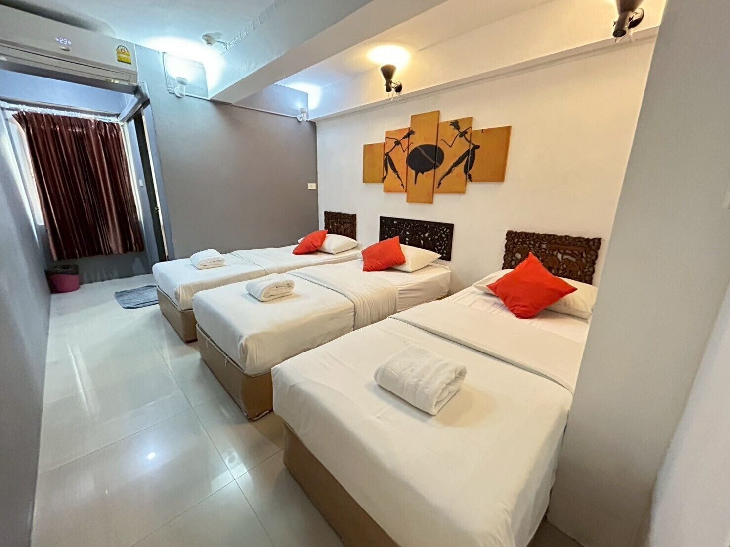 A hotel room with three single beds, each with white linens and red pillows. There is a wall-mounted artwork above the beds, and a ceiling light fixture.