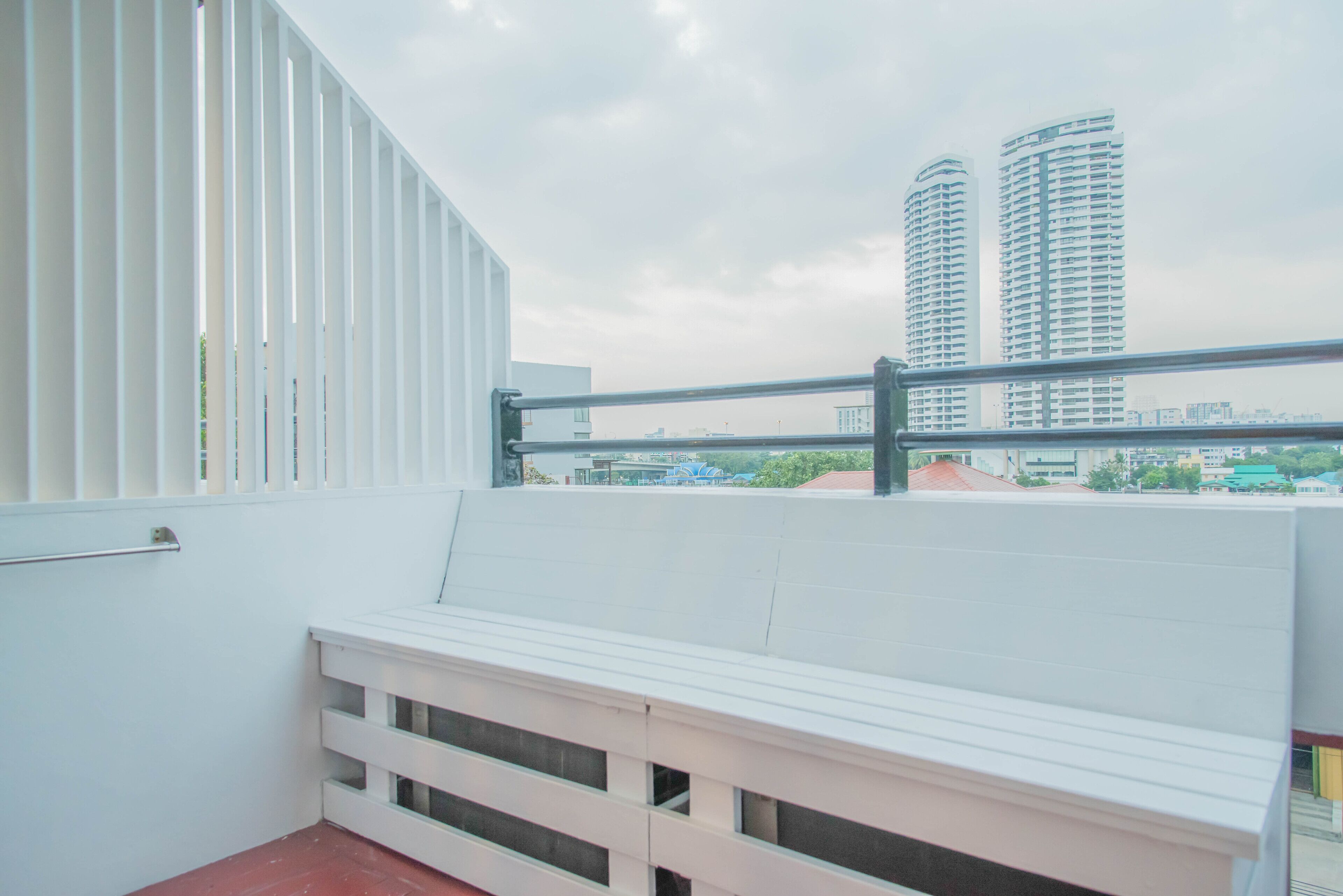 A balcony with white railings and a bench, overlooking tall buildings and a cloudy sky.