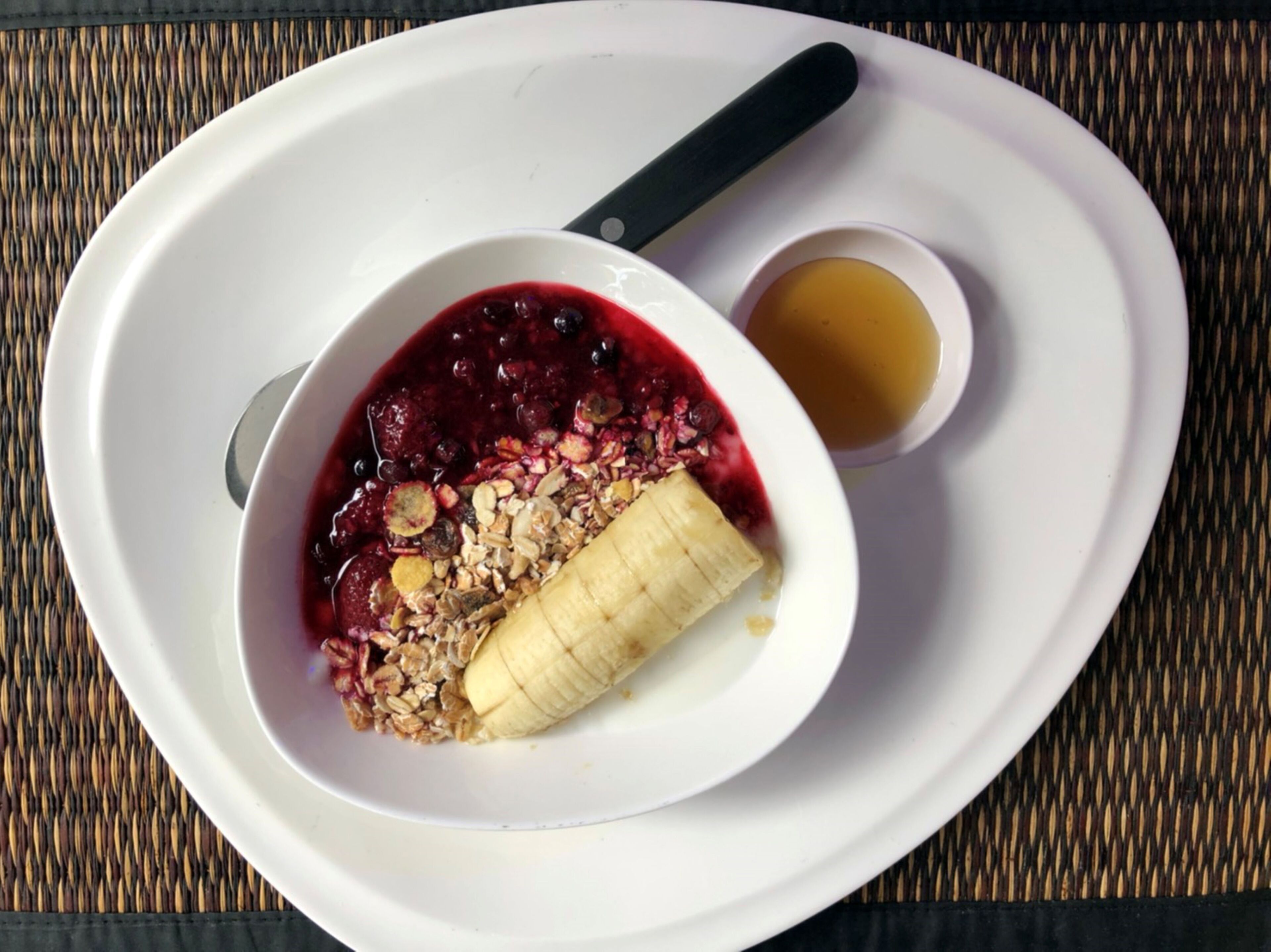 A white plate with a bowl of granola and berries, a banana, and a small cup of honey.