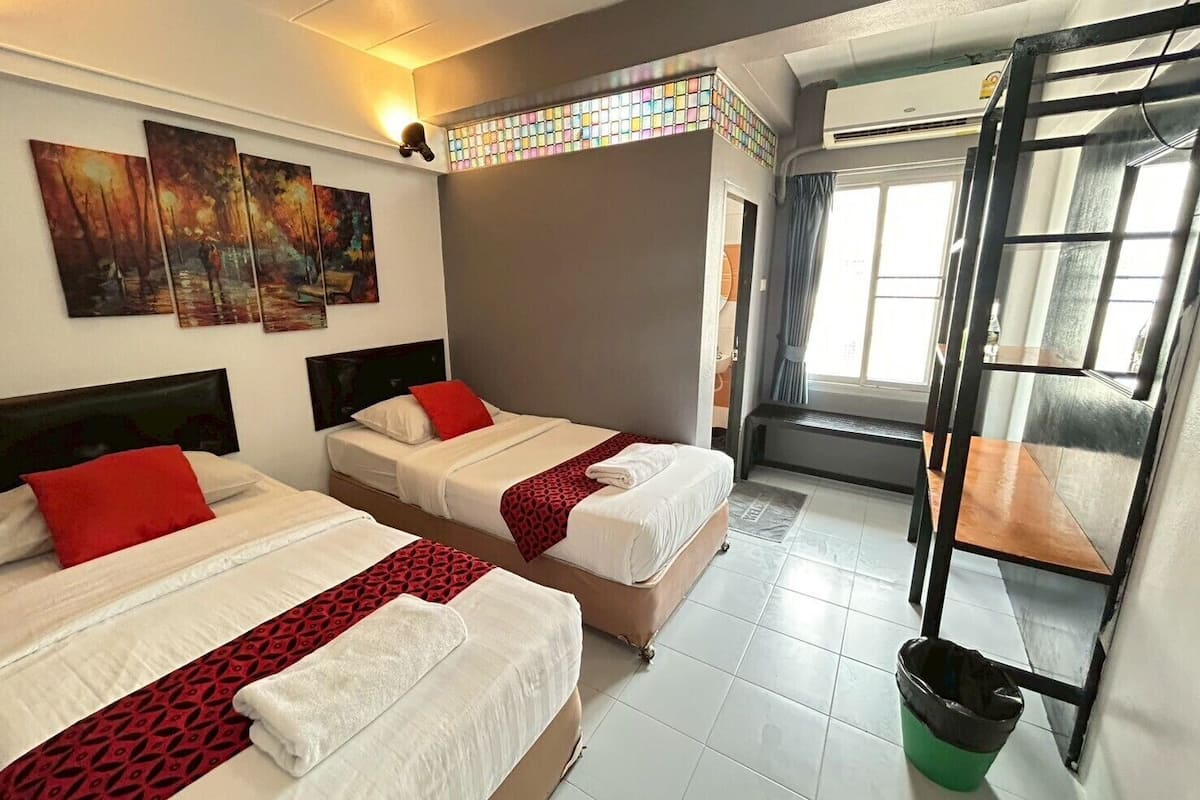 Une chambre d’hôtel avec deux lits, un lit superposé, une fenêtre avec des rideaux, une poubelle et des œuvres d’art au mur.