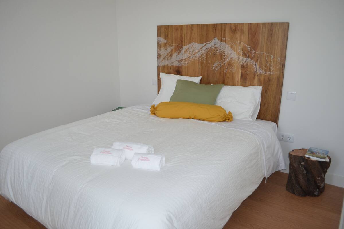 Een net opgemaakt bed met wit beddengoed, een houten hoofdbord met een bergmotief, en een klein houten krukje met een boek ernaast.