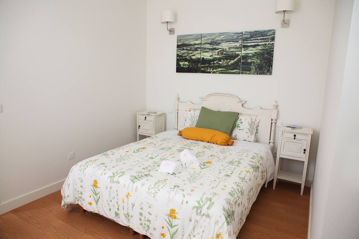 Een netjes opgemaakt bed met bloemenbeddels, een groen kussen en twee witte nachtkastjes met lampen.