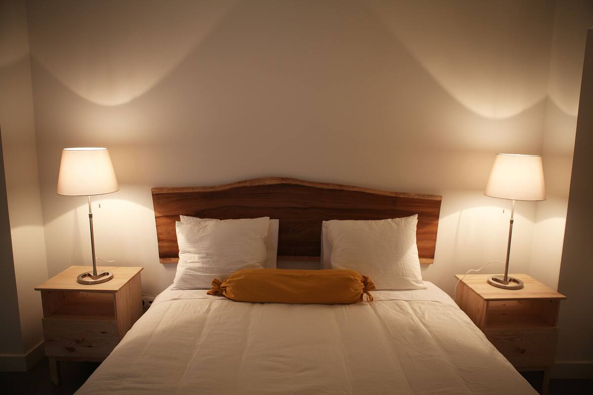 Een net opgemaakt bed met witte lakens, een houten hoofdbord en twee nachtkastjes met lampen.