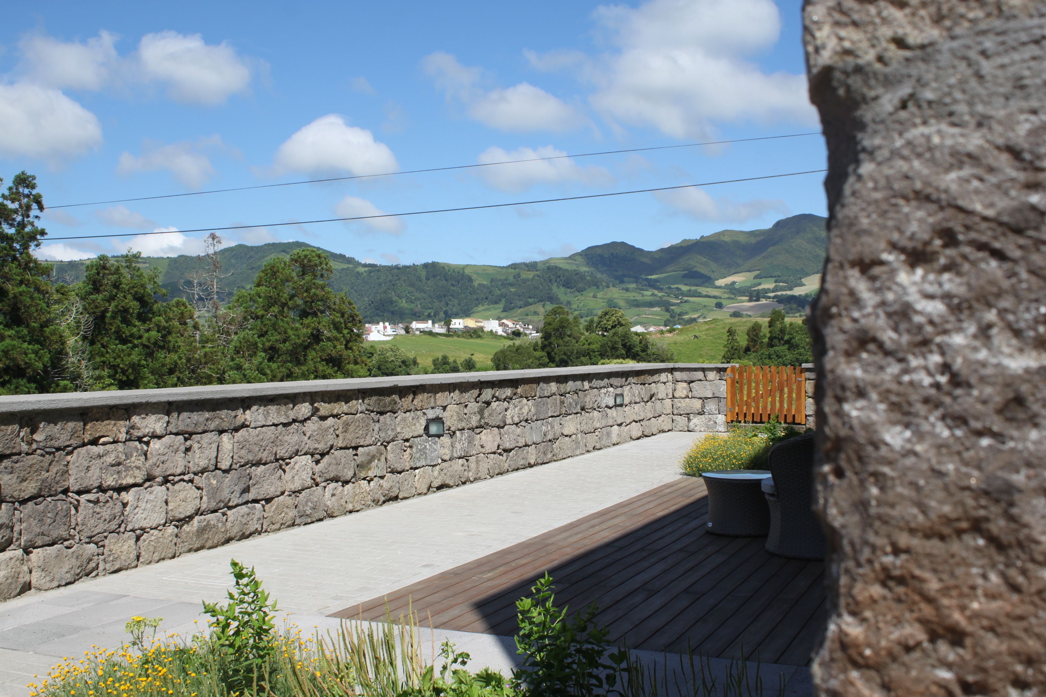 Un mur de pierre avec une terrasse en bois, des sièges d’extérieur et une vue sur des collines verdoyantes et des bâtiments au loin.