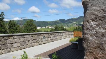 Un mur de pierre avec une terrasse en bois, des sièges d’extérieur et une vue sur des collines verdoyantes et des bâtiments au loin.