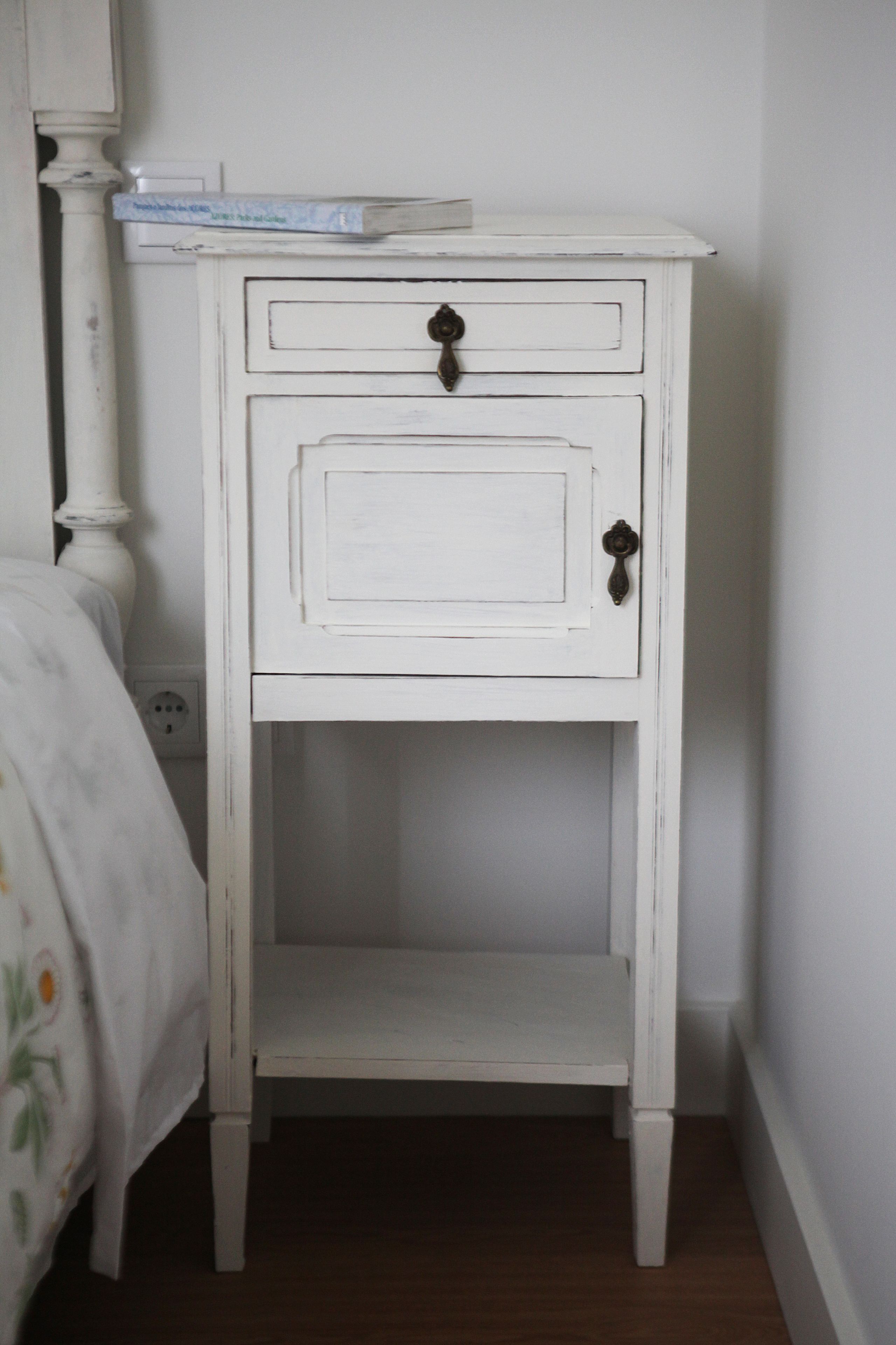 A white bedside table with a drawer and a shelf, positioned next to a bed with a floral bedspread.