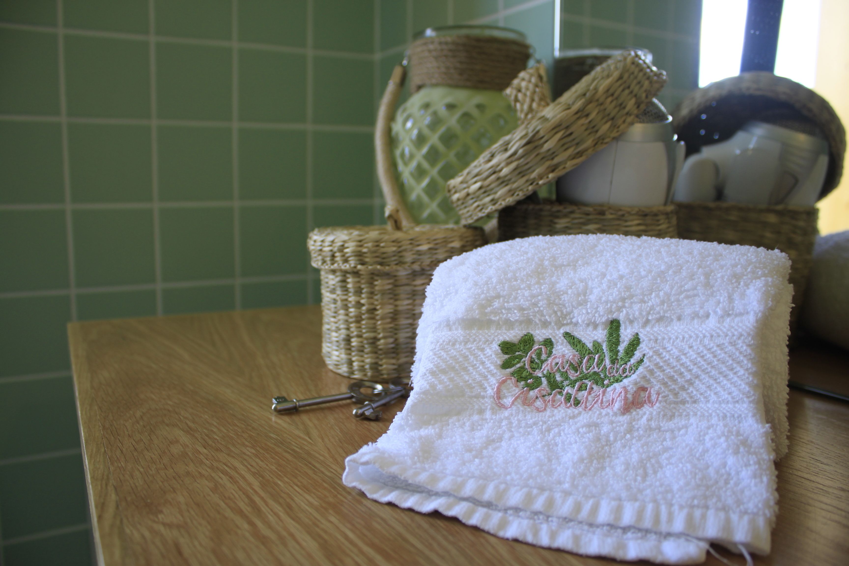 Un baño con tocador de madera, una toalla blanca con texto bordado, una cesta de mimbre y una pared de azulejos verdes.