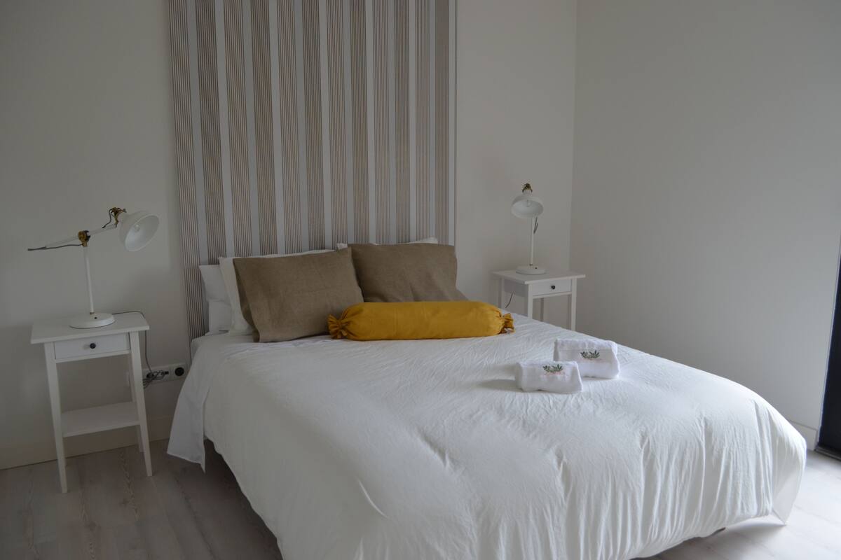 Een net opgemaakt bed met witte beddengoed en één geel kussen, zijkanten met twee nachtkastjes en lampen, in een kamer met een gestreept behang en een groot raam.