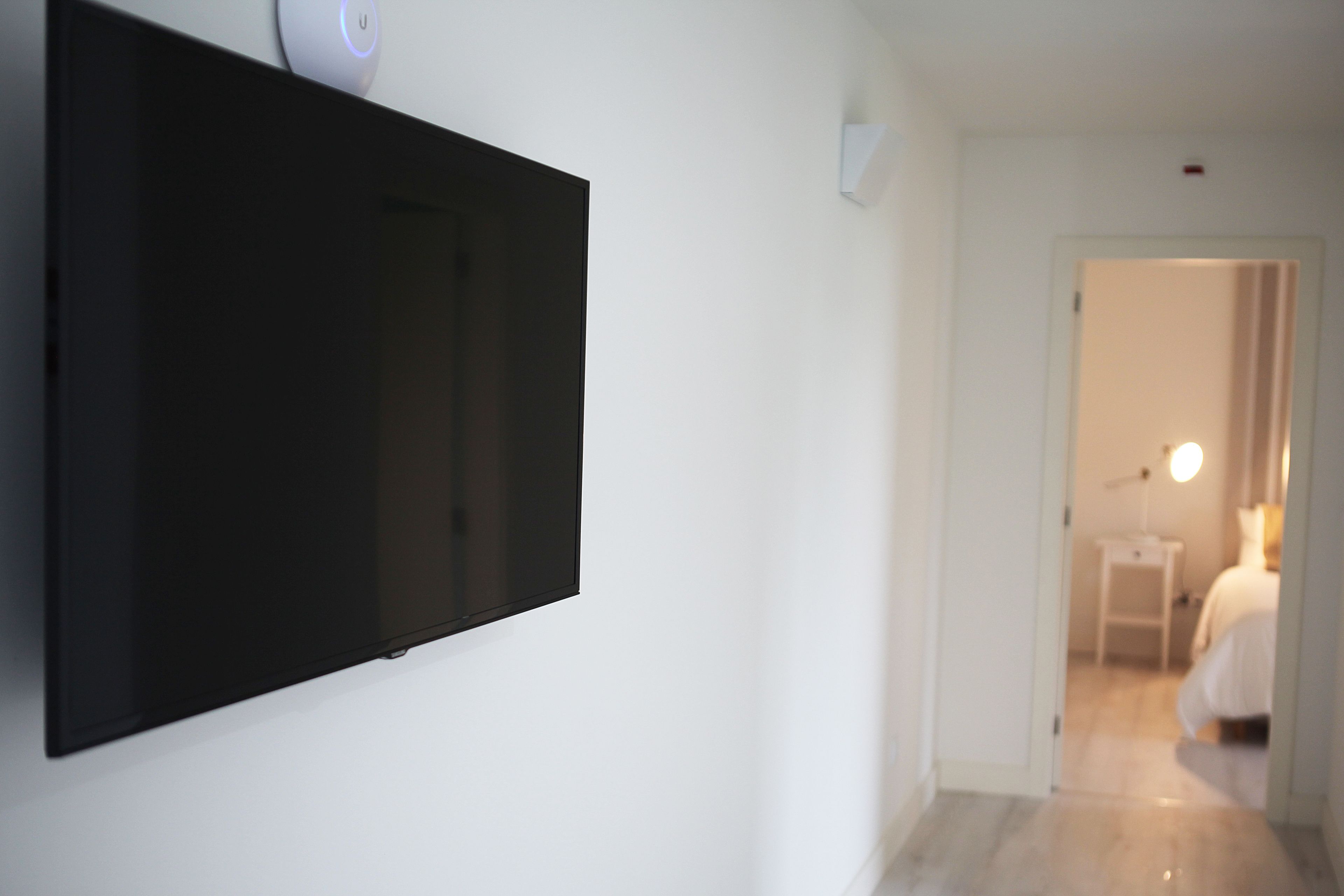 Habitación de hotel con televisor de pantalla plana en la pared, una mesita de noche con lámpara y cama con ropa blanca.