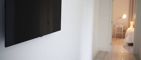 Habitación de hotel con televisor de pantalla plana en la pared, una mesita de noche con lámpara y cama con ropa blanca.