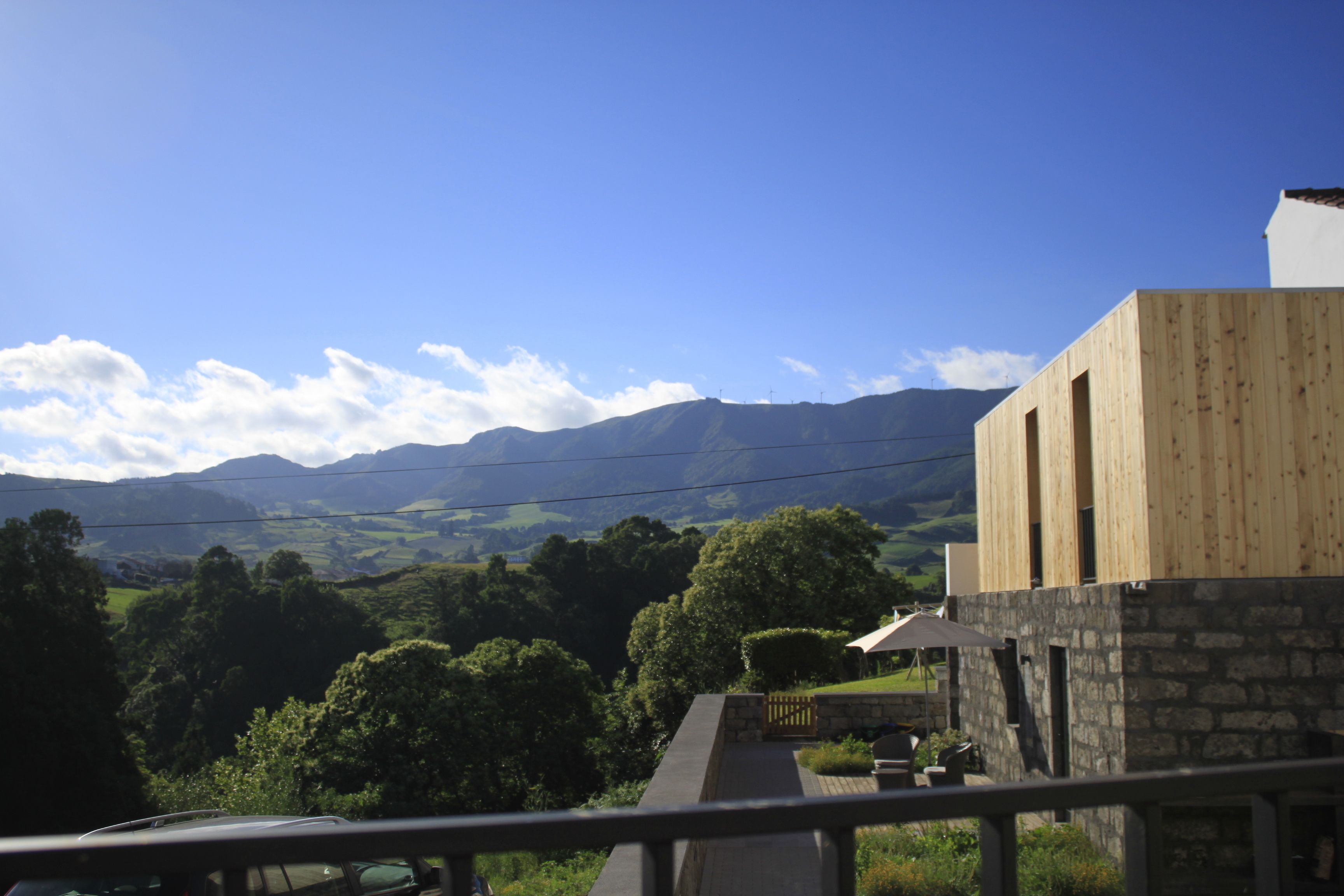 Un bâtiment moderne à la façade en pierre et au revêtement en bois, qui se détache sur un paysage de collines verdoyantes et d’un ciel bleu azur.