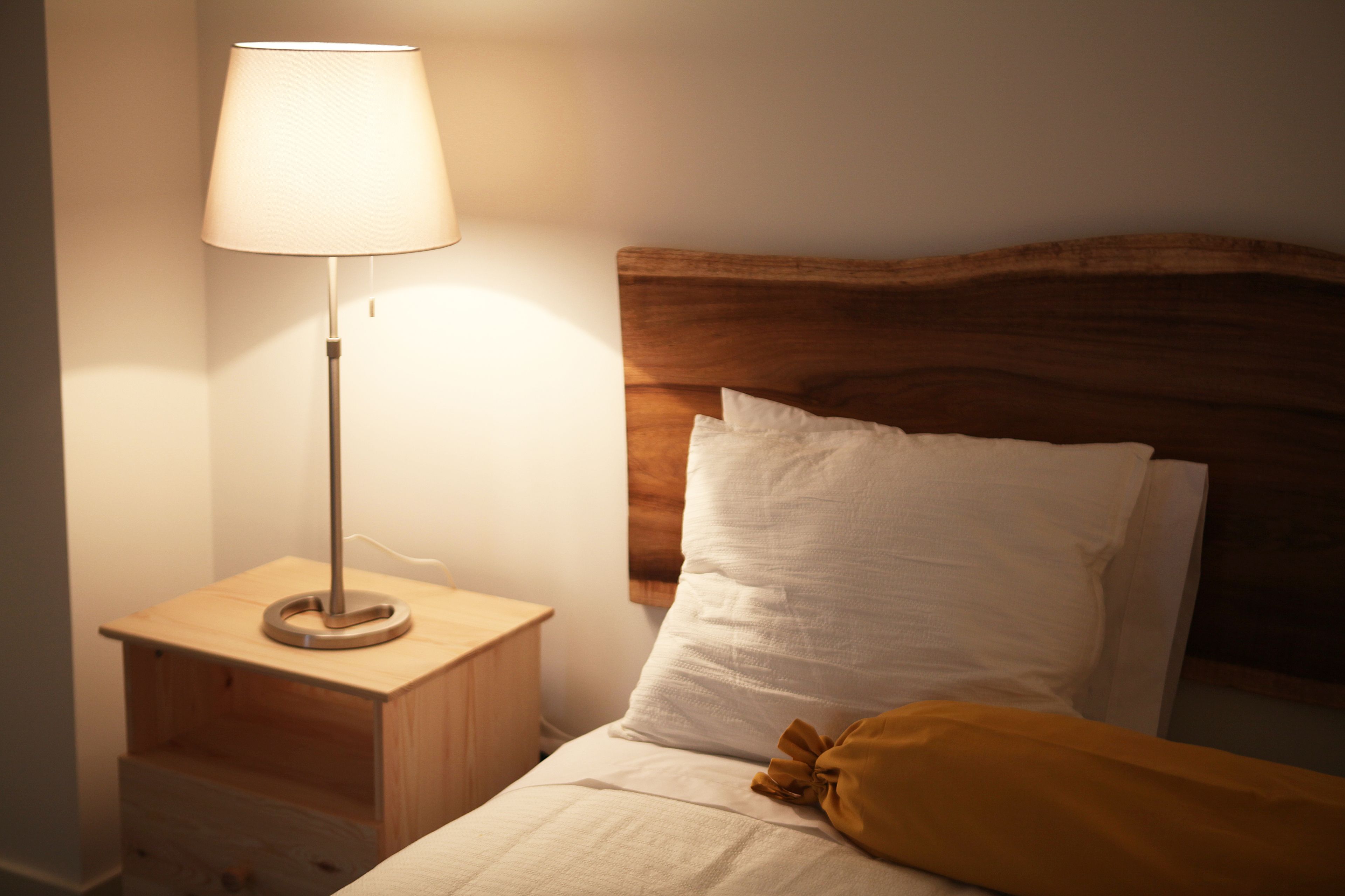 Un lit bien fait, avec du linge de lit blanc et une tête de lit en bois, une lampe de chevet et une table de chevet en bois dotée d’un tiroir.