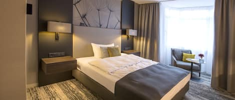 Una habitación de hotel moderna con una cama grande, lámparas de noche, una mesita con un florero y vistas al exterior a través de una ventana.