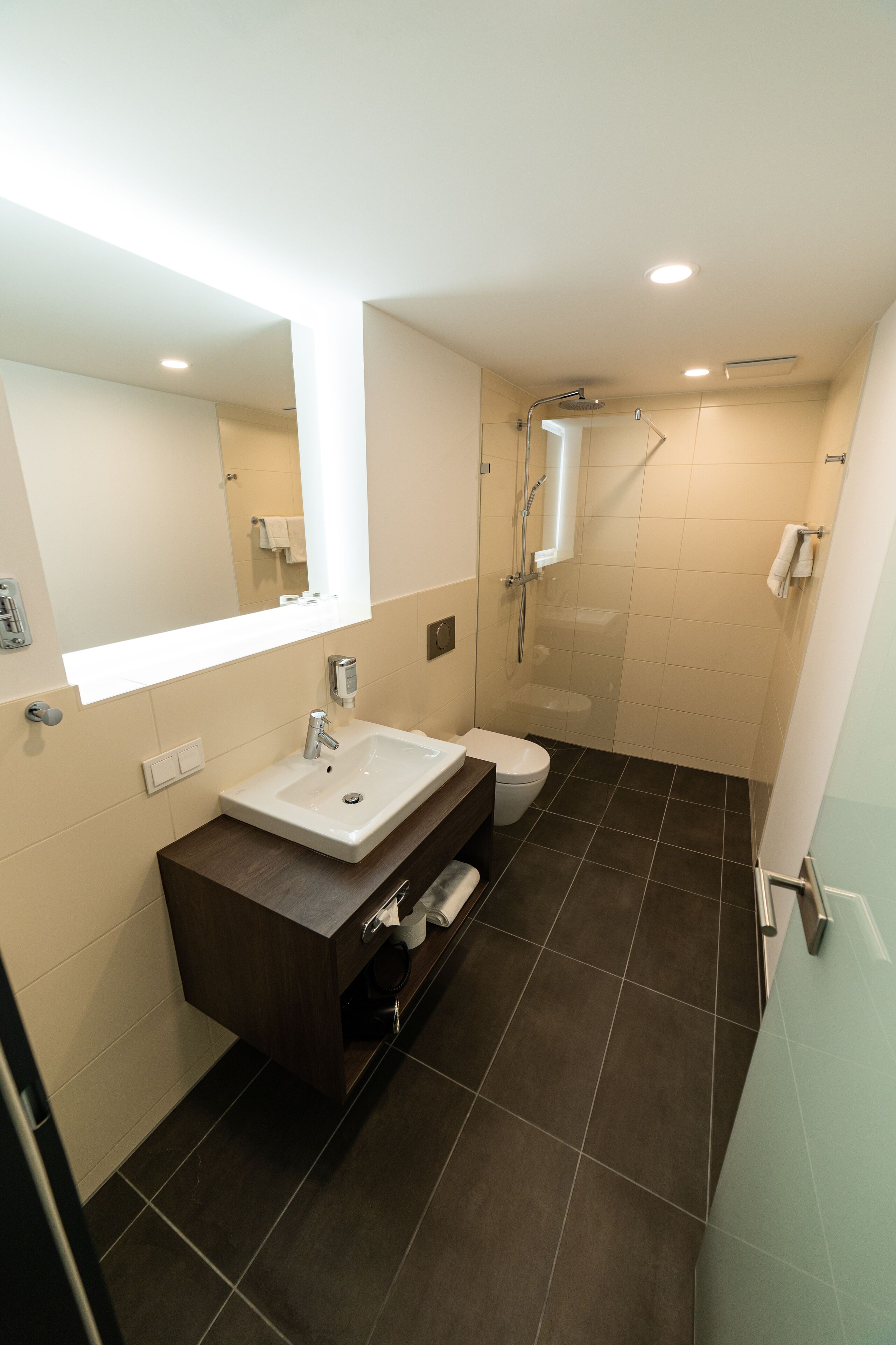 A modern bathroom with a white sink, dark brown vanity, a glass shower enclosure, and a white toilet.