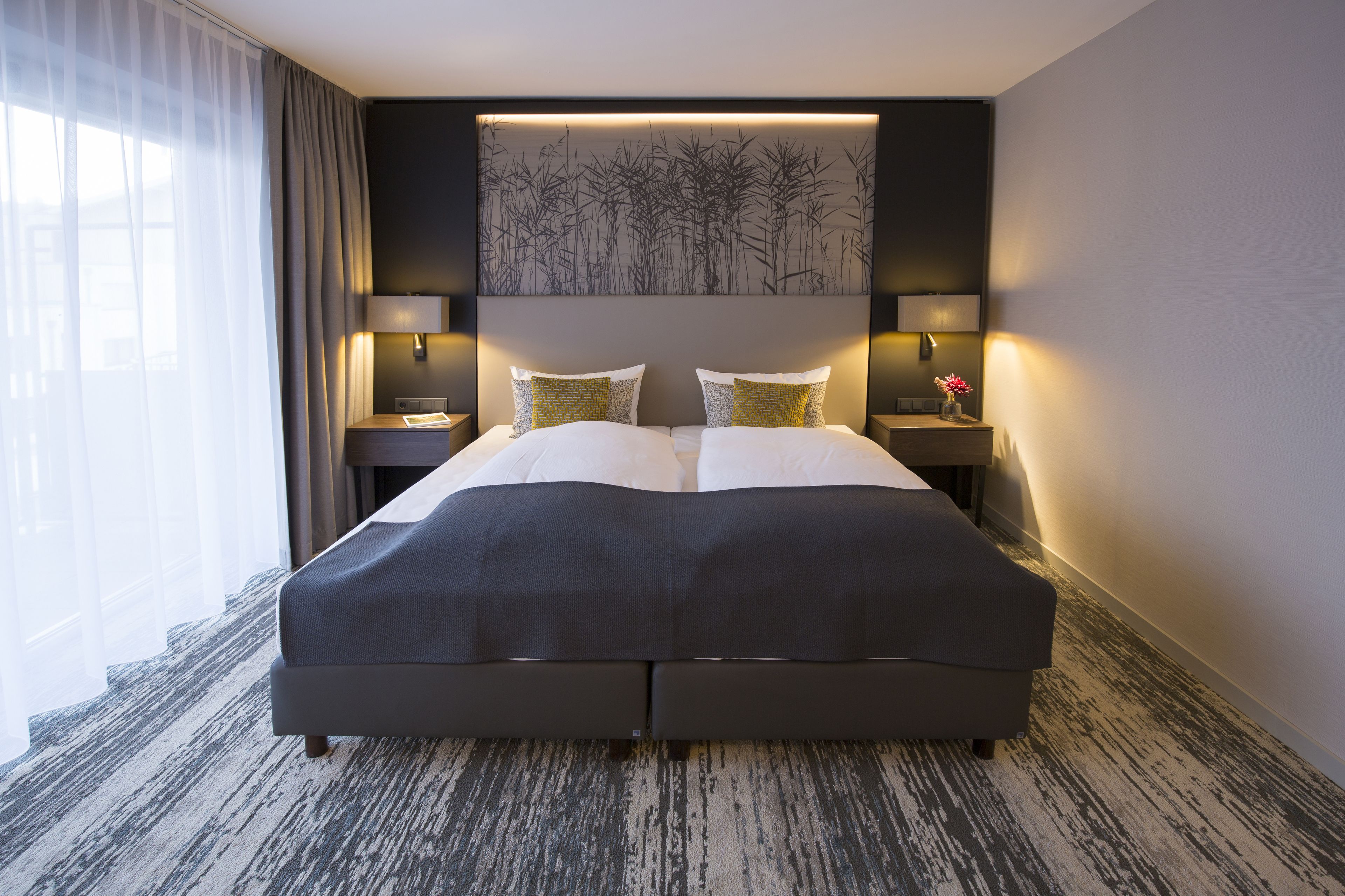 A large bed with a dark headboard, two bedside tables with lamps, a wall-mounted artwork, and a view of the outdoors through sheer curtains.