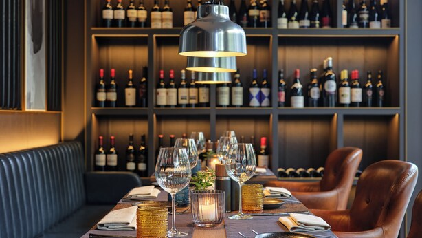 A restaurant setting with a long table, multiple wine glasses, and a wine bottle shelf in the background. - Kranz Parkhotel (Siegburg)
