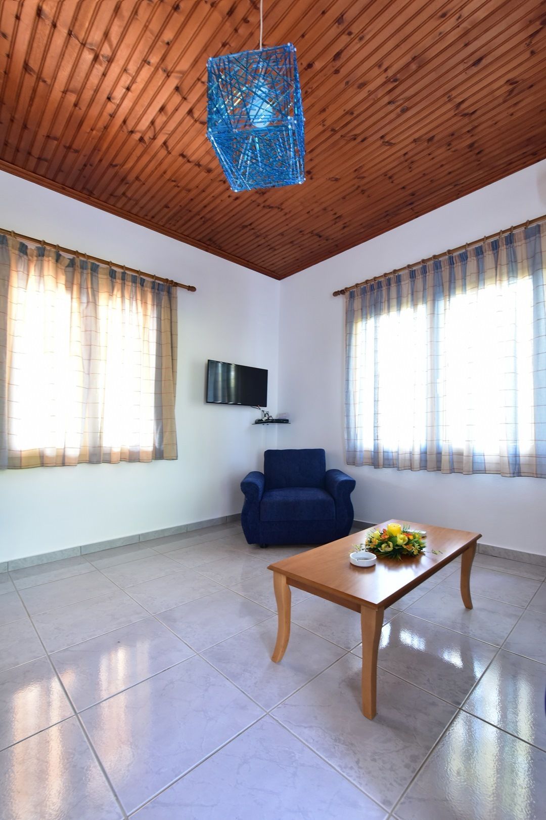 Une pièce spacieuse avec un plafond en bois, deux fenêtres avec des rideaux, un fauteuil bleu, une table en bois agrémentée d’un arrangement floral et un téléviseur à écran plat fixé au mur.