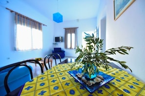 A dining area with a yellow tablecloth featuring blue floral patterns, a blue vase with green leaves, and a plate with shells. - Mouzakis Villas (Samos)
