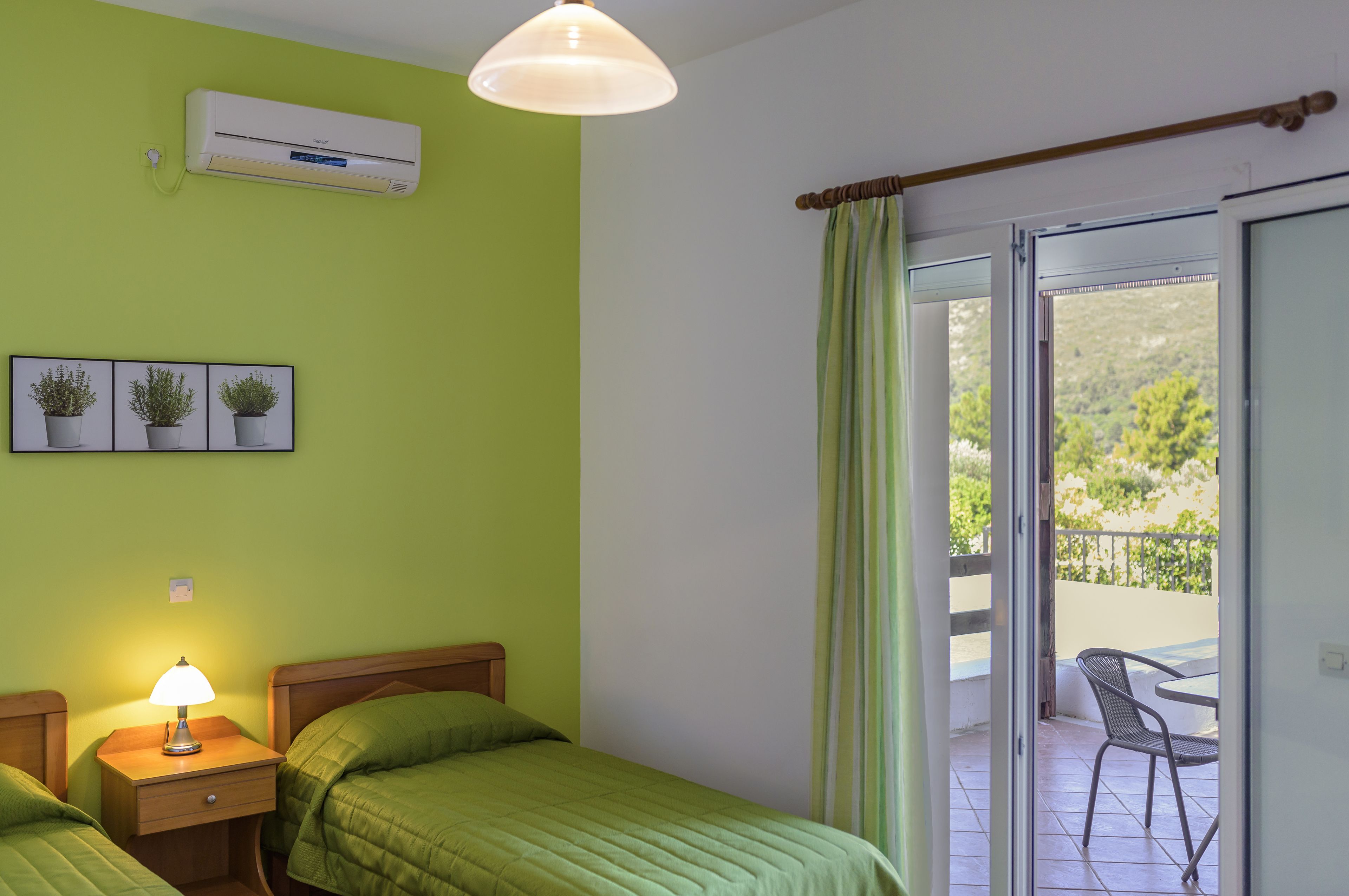 Una habitación con dos camas, una mesita de noche con lámpara, aire acondicionado y vistas a un balcón con mesa y sillas.