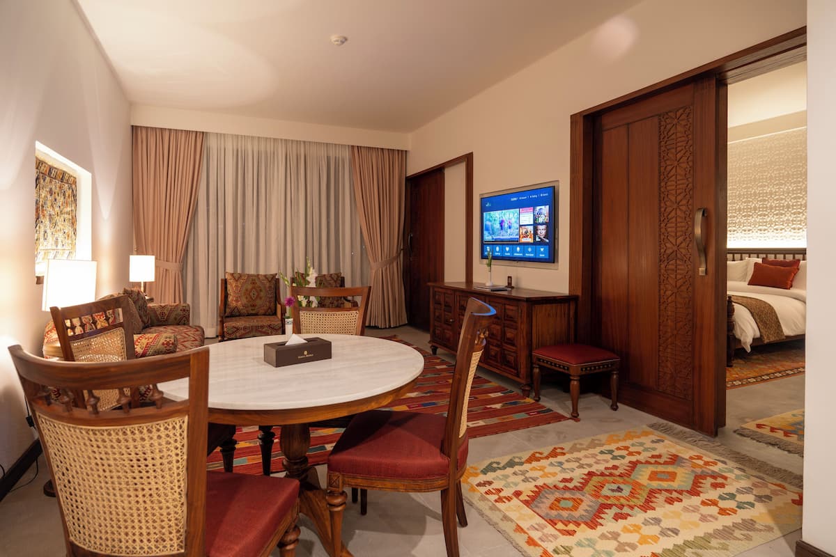 A hotel room with a round dining table, multiple chairs, a television mounted on the wall, and a bed with a patterned bedspread.