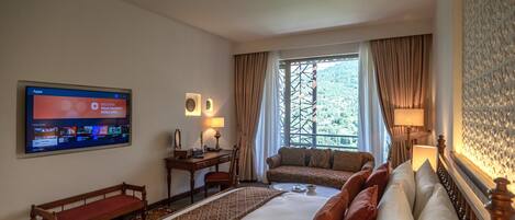 A hotel room with a large bed, a desk with a chair, a sofa, a TV mounted on the wall, and a window with curtains.