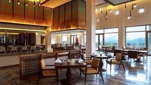 Un amplio hall de hotel con techos altos, grandes ventanales con vistas a la montaña y diversas opciones de asientos, incluyendo mesas y sillas.