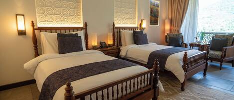 A hotel room with two wooden beds, a patterned wall, a balcony with a view, and a small table with a lamp.