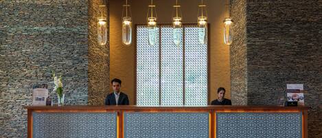A reception desk with a patterned backdrop, two individuals seated behind it, and hanging pendant lights.