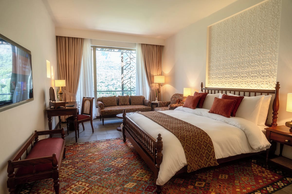 A hotel room with a large bed, a sofa, a desk, and a chair. There is a window with curtains and a view of greenery outside.