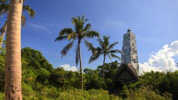 Una torre alta y esbelta con una estructura de techo de paja en su base, rodeada de densa vegetación tropical y palmeras bajo un cielo azul despejado.