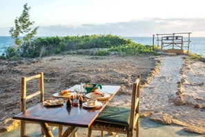 Une table en bois, dressée avec des assiettes, des bols et des verres, est placée sur une plage de sable, offrant une vue sur la mer et sur une structure en bois en arrière-plan.