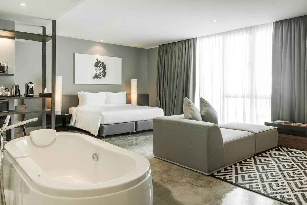 A modern hotel room with a large bed, a sofa, a bathtub, and a large window with curtains.