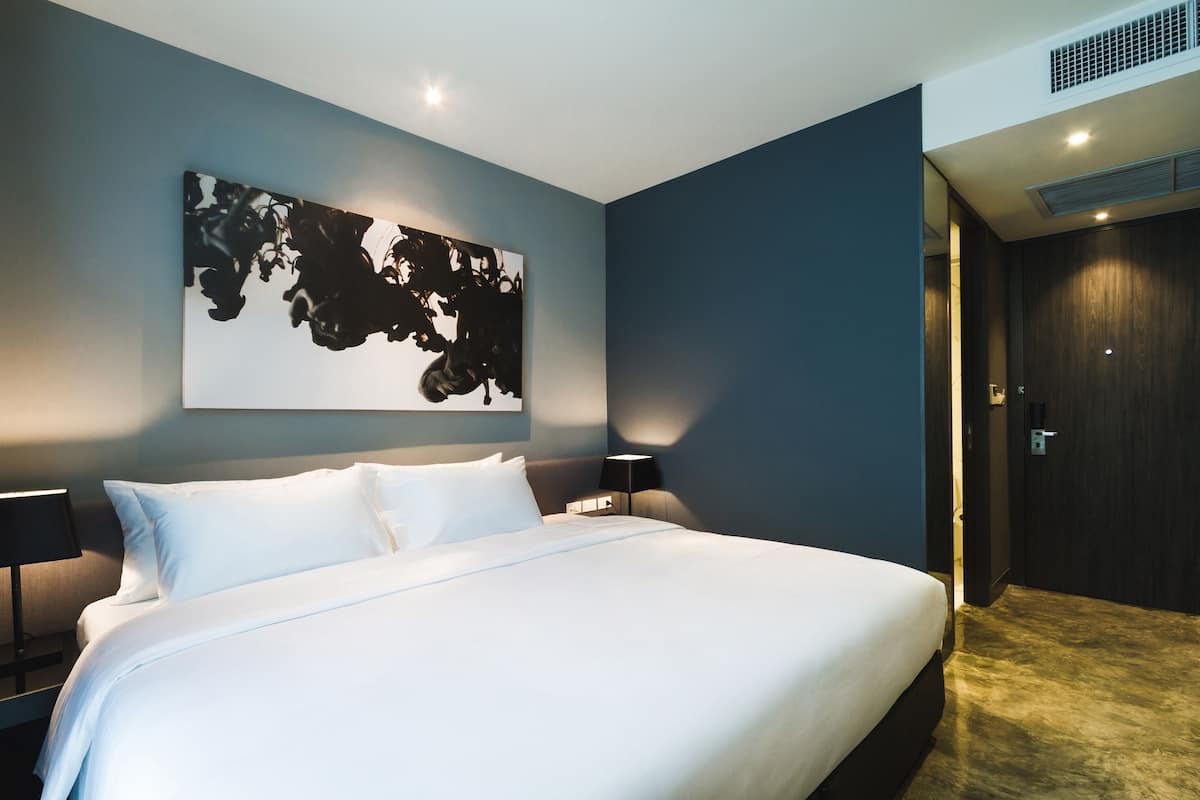 A modern hotel room with a large bed, a wall-mounted abstract artwork, a dark wooden door, and a nightstand with a lamp.