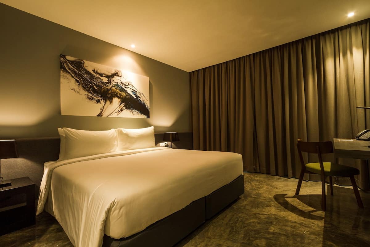 A hotel room with a large bed, a desk with a chair, a painting on the wall, and a curtain covering the window.